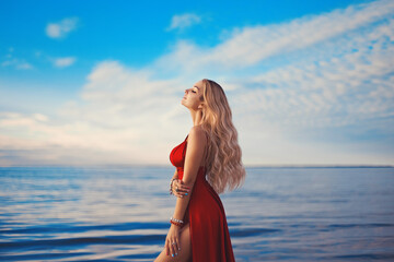 A gorgeous blonde girl in a sexy red dress with deep slits on the hips against the background of a sandy beach, blue sea and a sunset sky.