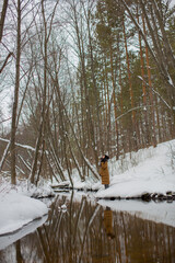 A girl in a brown jacket stands near the river against the background of the winter forest looking up. Winter forest. Winter river. High quality photo