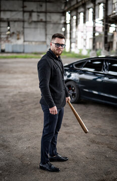 Brutal strong guy in jeans holding a baseball bat. Brutal image. Bearded man in sunglasses. Fashion car on the background.