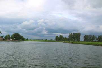 Wide angle shot of a landscape with a water body, crop fields, and cloudy blue sky.