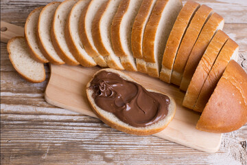 A loaf of bread and a slice of bread spread with chocolate-nut cream on a wooden background. Delicious Breakfast with chocolate and nut paste.