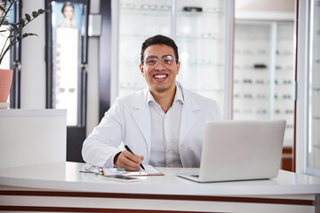 Smiling bespectacled optometrist seated at the table in his office - obrazy, fototapety, plakaty