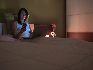 Young woman working on her laptop and looking at social networks on the smartphone in bed before going to sleep.
