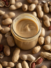 Homemade peanut butter in a glass jar with peanuts in shells and peeled scattered around on a brown craft background in natural light