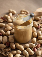 Homemade peanut butter in a glass jar with peanuts in shells and peeled scattered around on a brown craft background in natural light