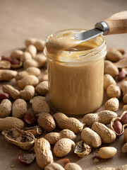 Homemade peanut butter in a glass jar with peanuts in shells and peeled scattered around on a brown craft background in natural light