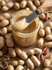 Homemade peanut butter in a glass jar with peanuts in shells and peeled scattered around on a brown craft background in natural light