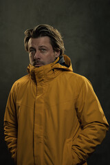 Blonde man in yellow coat in front of grey wall.