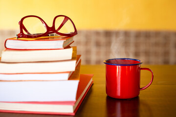 Pilha de livros com óculos vermelho em cima e uma caneca vermelha com café quente sobre a mesa.