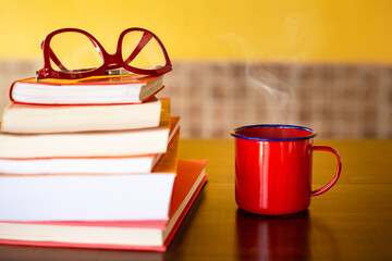 Pilha de livros com óculos vermelho em cima e uma caneca vermelha com café quente sobre a mesa.