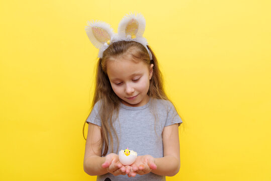 Cute little child wearing bunny ears on Easter day. Girl holding a painted egg on light yellow background. Studio photography