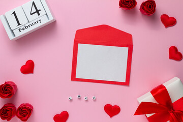 The concept of Valentine's Day. Envelope with a blank sheet, hearts, wooden calendar gift with a red ribbon and roses on a pink background. Flat lay, copy space.