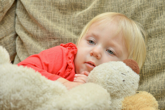 A little girl lies with a toy dog on the bed and looks to the side.