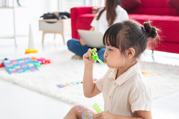 Cute girl standing in bedroom by the bed at home and blowing soap bubbles. Adorable girl blowing soap bubbles in bedroom. Young women at home with little girl are having fun and blowing soap bubbles.