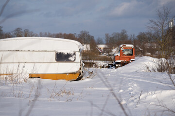 An old car trailer for travel is presented in the deep snow and behind it you can see a red tractor in the distance