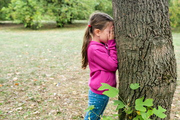 Children playing hide and seek in the park on an autumn day. The girl standing near a tree and covering her face with hands. Outdoor activities with kids