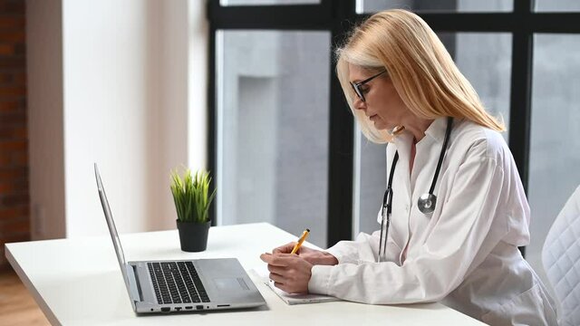 Confident and helpful middle-aged MD, pediatric, physician wearing white medical gown in modern clinic office and talks with patient via video connection on the laptop, consults online. Side view