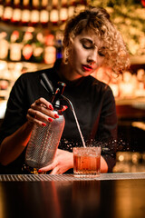 attractive female bartender stands behind the bar and accurate pours drink from siphon into glass