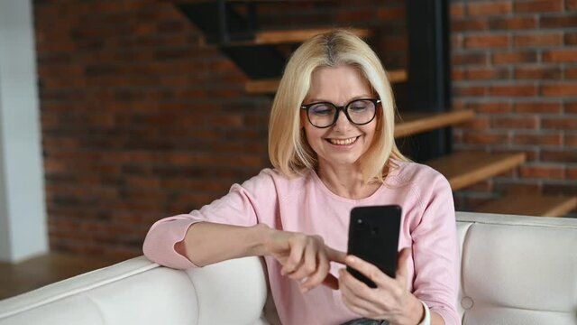 Happy smiling middle-aged woman holds smartphone sitting on the couch. Charming lady spends leisure time websurfing, shopping online, scrolling news feed on the phone at home