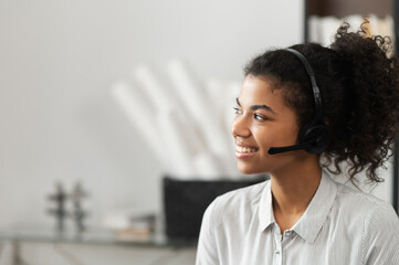 Young thoughtful African American female sudent in a headset is listening to an online webinar, studing remotely from home or working in the customer service department as a call center operator
