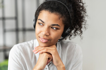 Fototapeta na wymiar Thoughtful African American millennial woman in a headset is listening to an online webinar, sitting with her hands on a chin, working in the customer service department as a call center operator