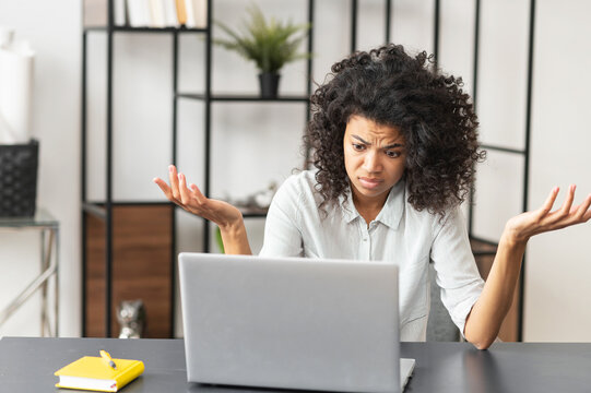 Young African American female office worker manager feeling stressed, tired, and overworked, does not know how to fix a problem, sitting at the desk in front of laptop, struggling and raising hands up