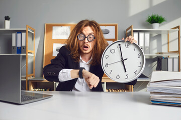 Employee working under pressure. Funny stressed young office worker with messy long hair holding clock, looking at wristwatch and panicking over missed deadline or being terribly late for work meeting - obrazy, fototapety, plakaty