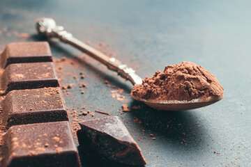 Brown cocoa powder in the spoon, chocolate bar, chopped chocolate cubes on dark background with copy space