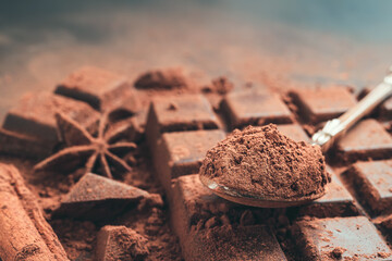 Brown cocoa powder in the spoon on chocolate bar, chopped chocolate cubes on dark background with copy space