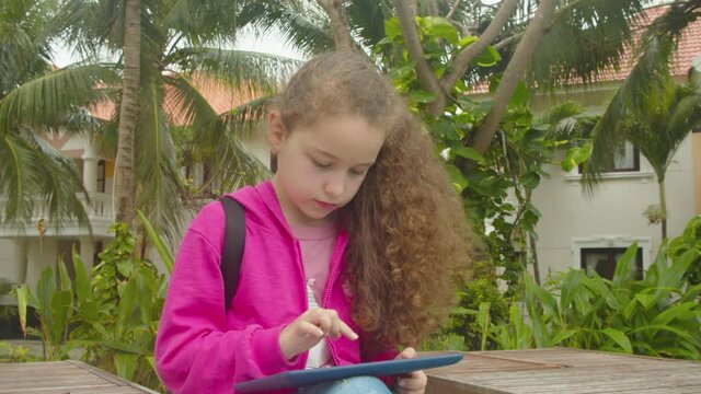 Cute little girl child using a digital tablet technology device telephone or laptop.Plays in the phone sitting on street alone.Children tech addiction concept play game.