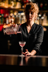 front view of female bartender pouring cocktail from mixing cup into crystal glass