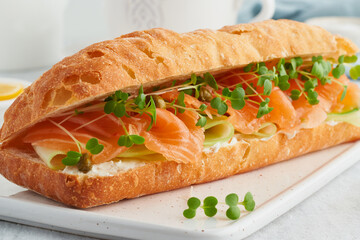 Big sandwiche with salmon, cream cheese, cucumber slices on white concrete table. Morning healthy breakfast with fish and cup of drink, side view, close up