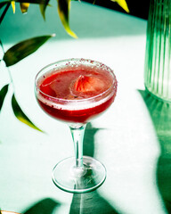 red tropical cocktail with ice in the form of a crystal on a background of foliage and shadows from sunlight.Green background