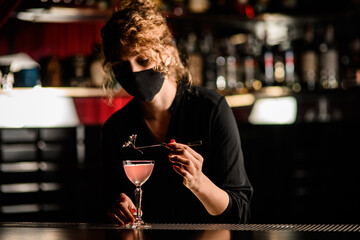 view of female bartender in mask that neatly decorates glass of cocktail with flower sprig