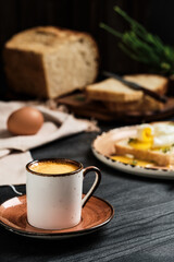 Espresso coffee close-up on black wooden table. Against blurred background, soft-boiled egg (poached) in slice of bread, covered with butter cream and herbs and loaf of sliced bread. Breakfast idea