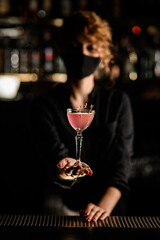 focus on beautiful glass of cocktail decorated with flower in the hands of woman in mask