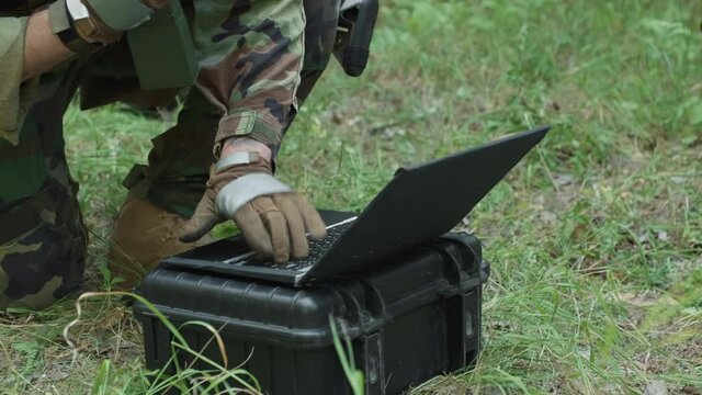 Tilting handheld closeup of special forces soldier entering data on laptop while talking via radio during important military operation in jungle