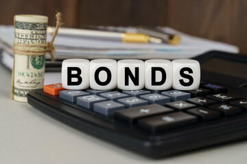 There are cubes on the calculator that say - BONDS. Nearby out of focus - dollars, notebook and pen