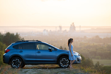 Happy young woman driver in blue dress enjoying warm summer evening standing beside her car. Travelling and vacation concept.