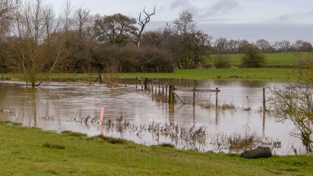 Fast running stream bursts its banks flooding farmland. Rural landscape image with reflections in water, partially submerged fencing. Distant views of tree lined countryside. England.