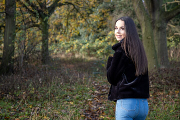 Naklejka na ściany i meble A young attractive woman with long brown hair in the winter time in a woodland park wearing a black coat