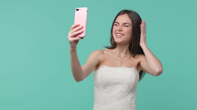 Cheerful bride young woman in white wedding dress posing isolated on blue turquoise background studio. Wedding concept. Doing selfie shot on mobile phone showing victory sign blowing sending air kiss