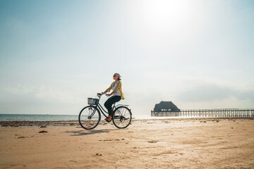 Fototapeta premium Young woman dressed light summer clothes riding old vintage bicycle with front basket on the lonely low tide ocean white sand coast on Kiwengwa beach on Zanzibar island, Tanzania.
