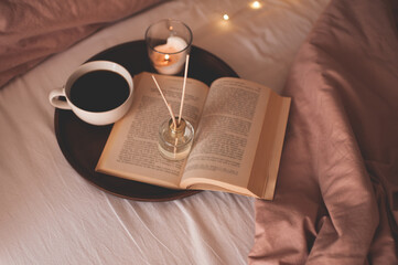 Cup of coffee with open paper book on wooden tray with liquid scented home fragrance in bed over...