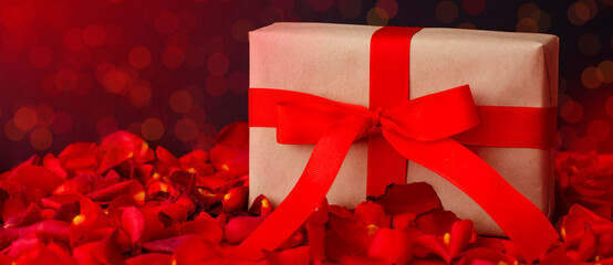 Gift box with a red bow ribbon and a red petals on a table for Valentine's day.