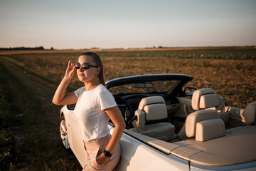 A beautiful young woman of European appearance on the face of sunglasses, she stands near her white convertible. Successful woman businesswoman and her white car.