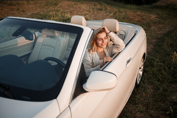 A glamorous luxury girl with blonde hair smiling while sitting on a white convertible. Young successful woman sitting in her white car
