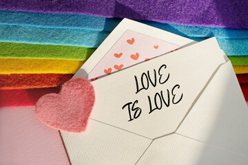 An open envelope with the words LOVE IS LOVE rests on a stack of fabric folded in the colors of the LGBT flag. Flat lay, top view. LGBT and Valentine's Day concept.