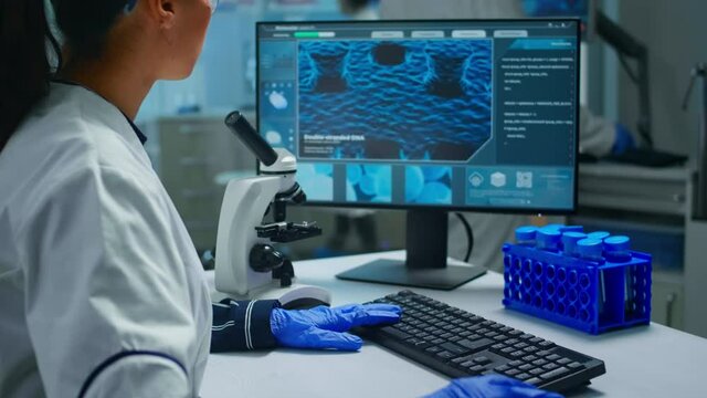 Biochemist using microscope and writing at computer sitting in equipped laboratory. Scientist doctor working with various bacteria, tissue, blood samples, pharmaceutical research for antibiotics