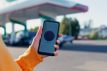 Woman holds a smartphone with a digital fuel meter on the screen in the background of a gas station and a car.